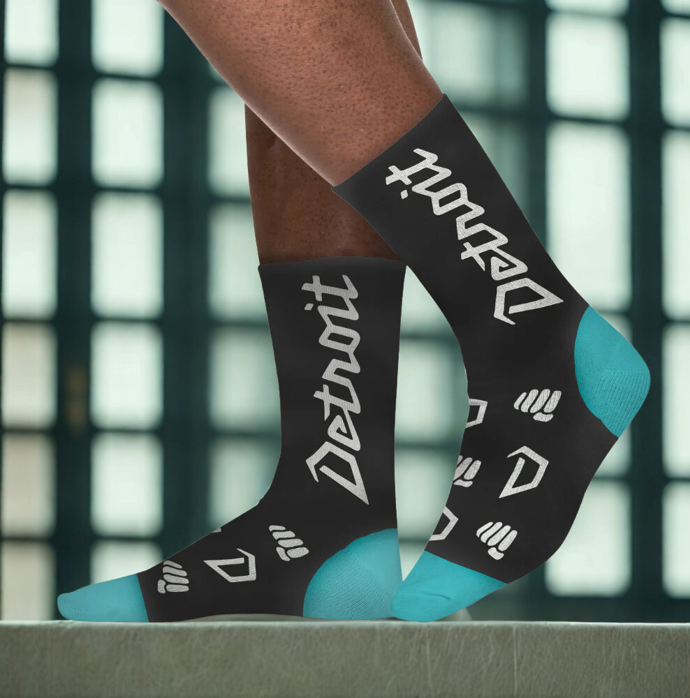 Black socks with 'Detroit' text and blue accents worn by a person against a blurred indoor background.